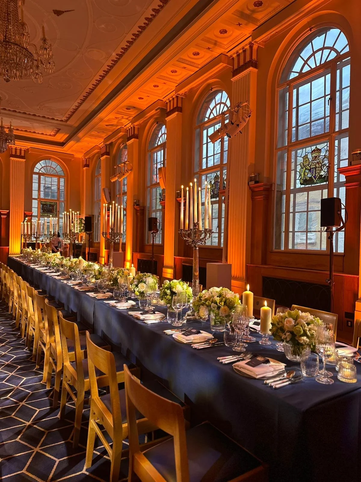 Elegant banquet hall with a long black table set for a formal event, decorated with white floral centerpieces, candles, and glassware, featuring tall windows, ornate ceilings, and wall sconces, illuminated by warm lighting.