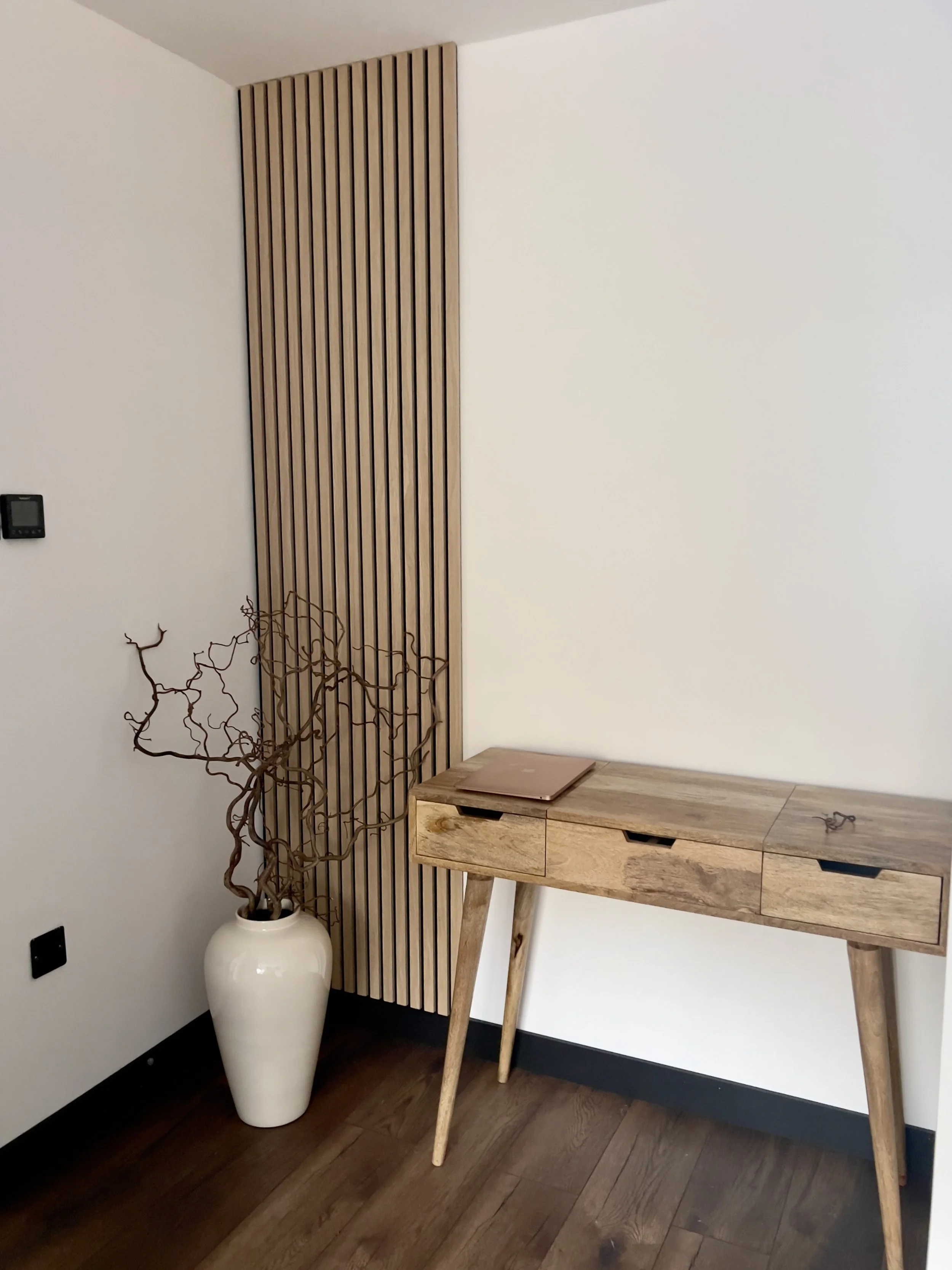 Interior corner of a room with a white wall, a wooden slat accent wall, a tall white vase with twisted branches, a wooden table with a notebook, and dark wood flooring.
