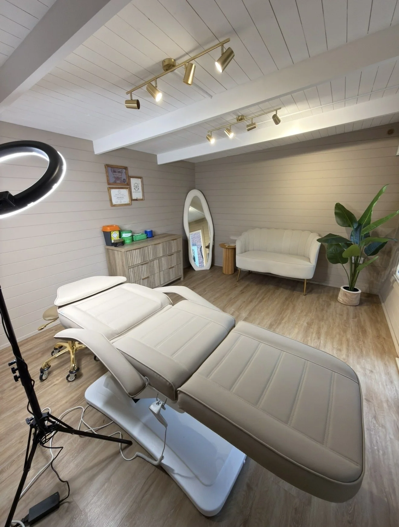 A room set up for a medical or spa treatment, featuring a reclining treatment chair with an adjustable headrest, a standing ring light, a mirror, a couch, a wooden floor, white walls with framed certificates, a dresser with containers, a large plant, and ceiling lights.