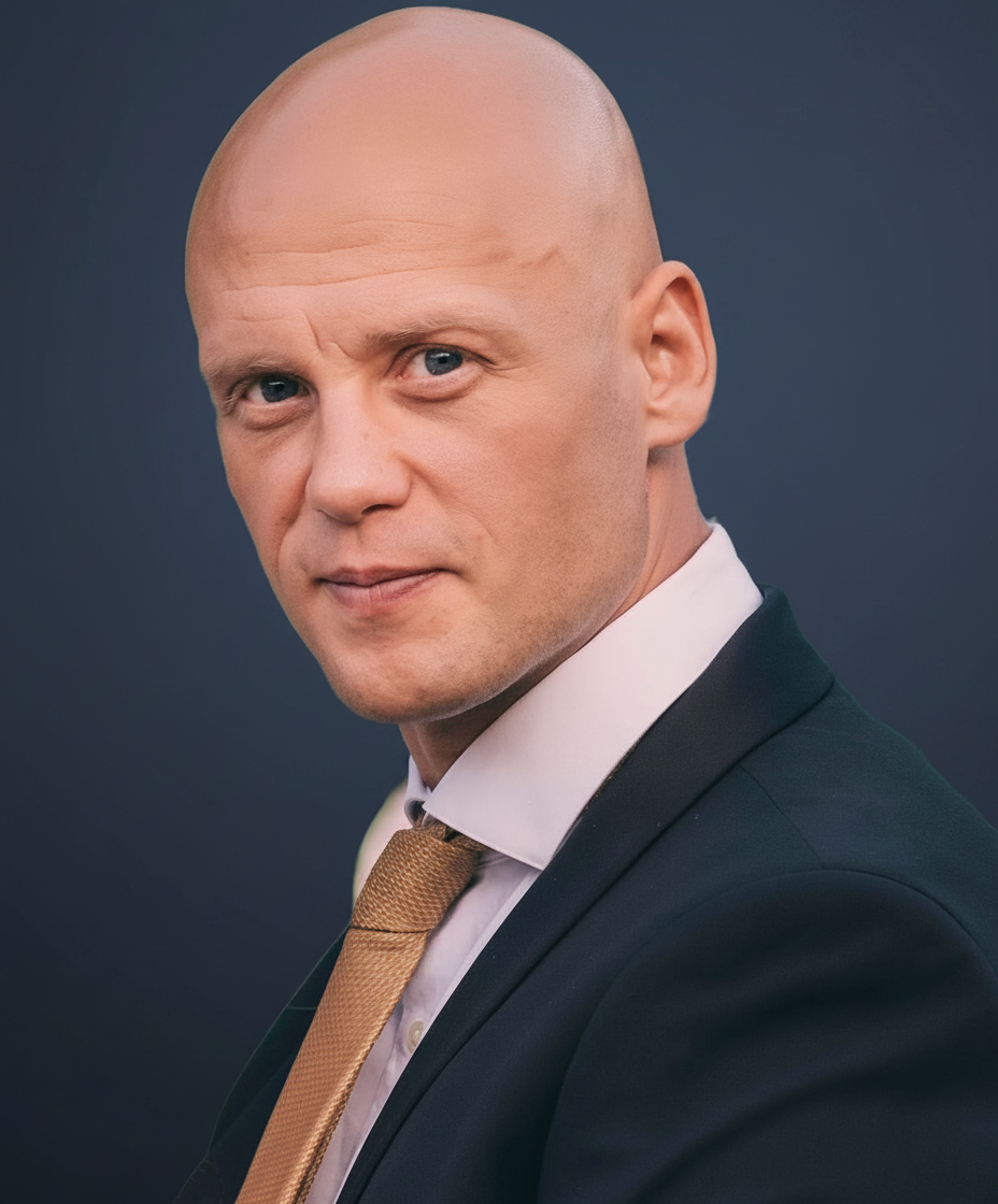 A bald man in a dark suit, white shirt, and brown tie looks into the camera with a serious expression against a dark background.