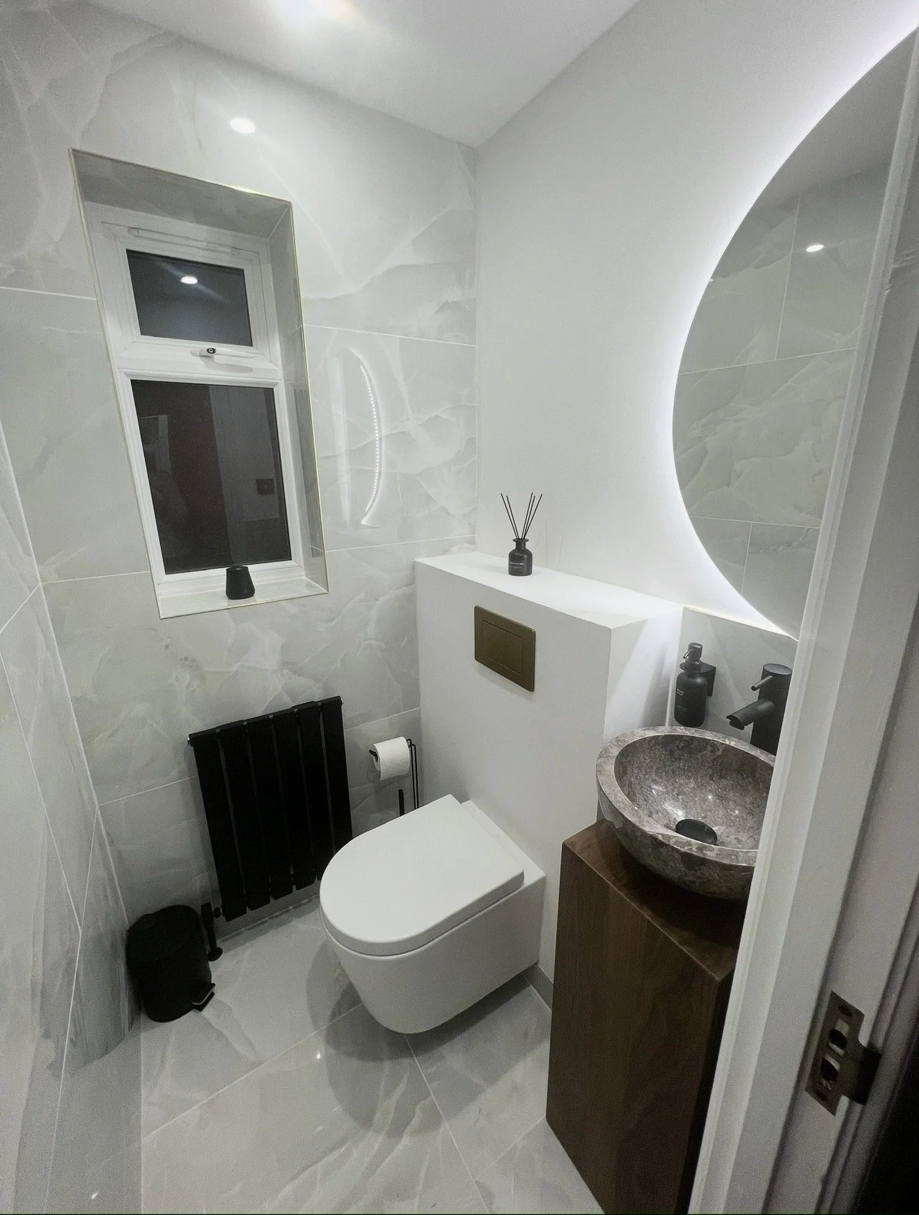 Modern small bathroom with white marble walls, a window, a white toilet, a black radiator, a small sink with a stone basin, a wooden cabinet, and black decor items.