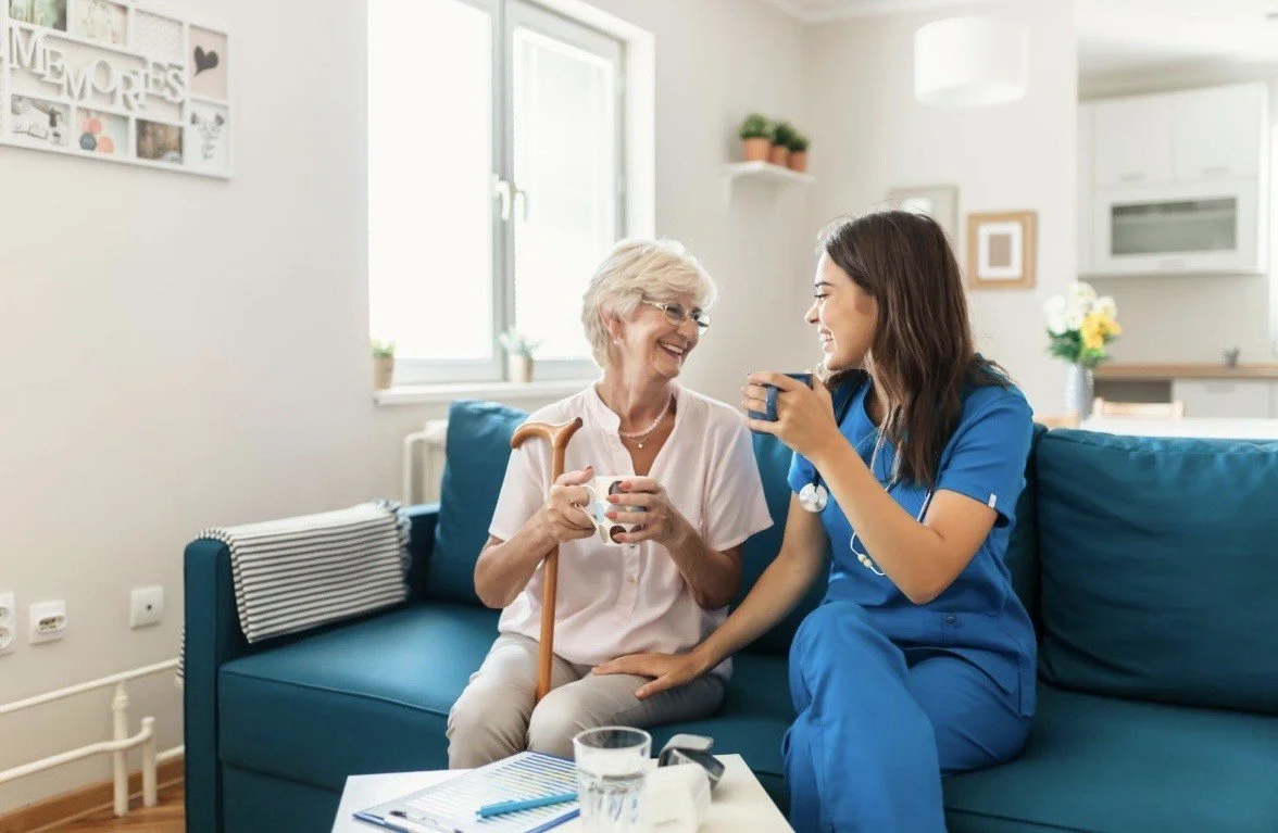 A nurse in blue scrubs and an elderly woman with a cane sitting on a blue sofa, sharing a joyful moment and cups of coffee in a bright, cozy living room.