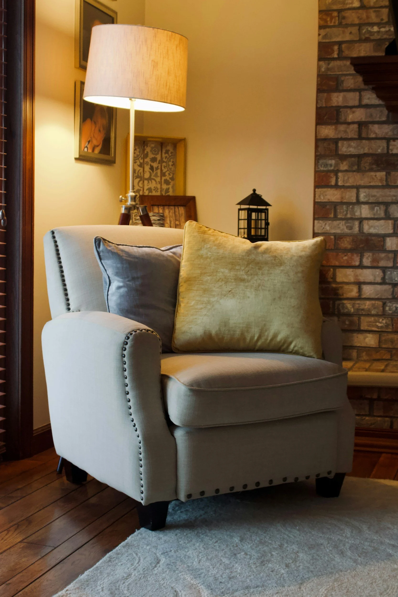 Close-up of a beige armchair with black wooden legs and silver nailhead trim, placed on a light-colored rug in a cozy living room with wooden floors, a brick fireplace wall, and a side table with various decorative items and pictures on the wall.