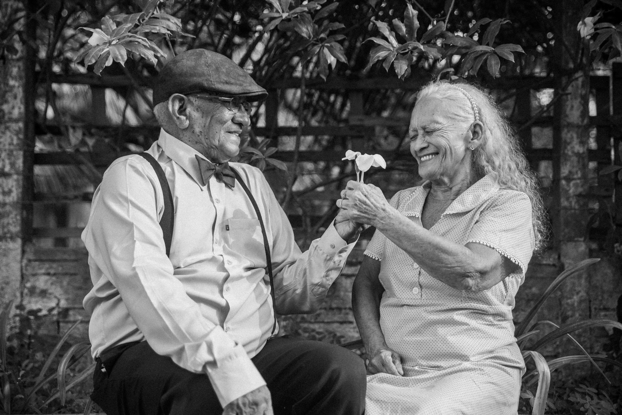 An elderly man and woman sitting outdoors surrounded by plants, smiling and exchanging a flower.