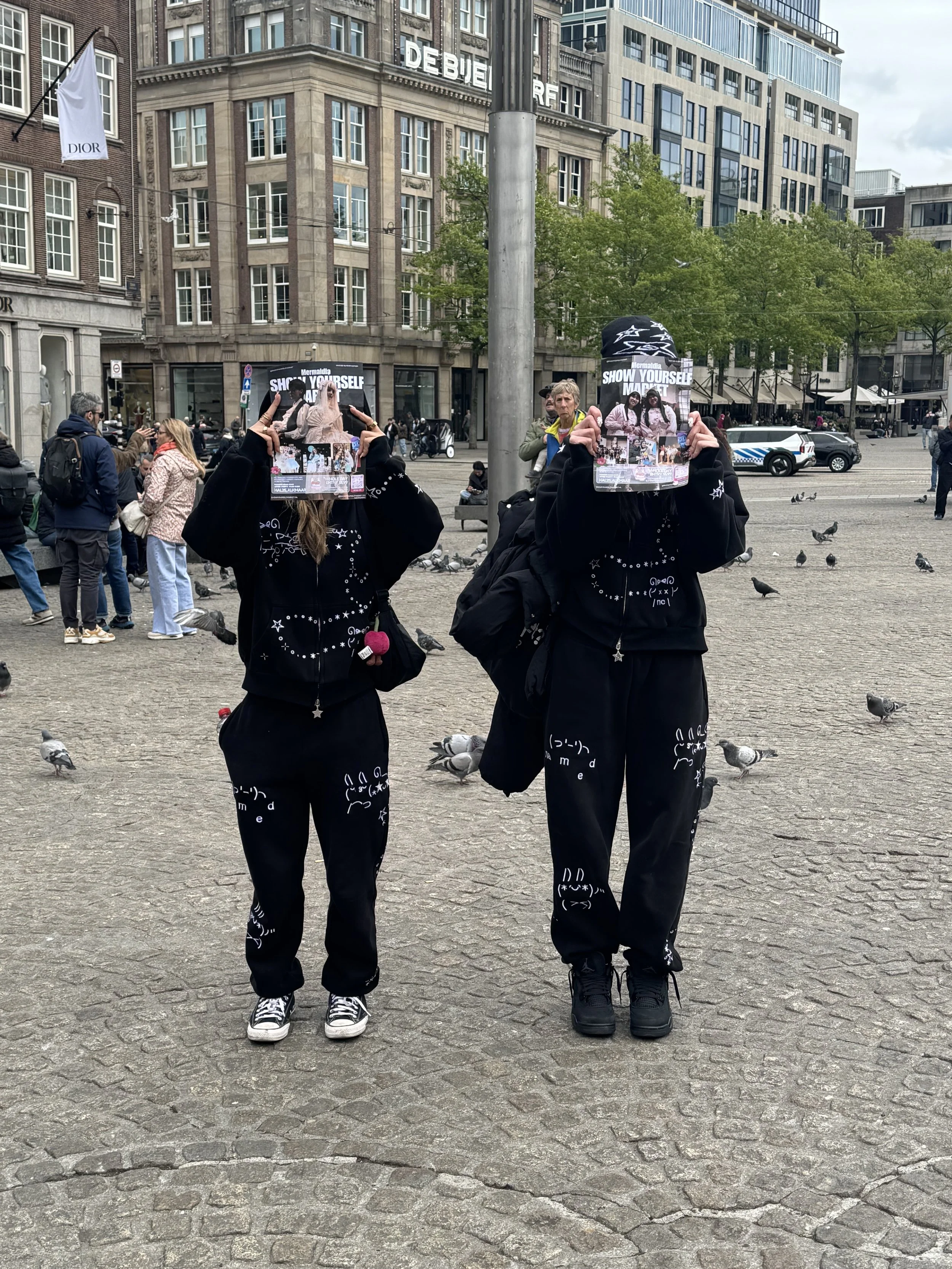 Twee mensen in zwart trainingspak met grafische witte patronen op een openbare plein, die poster vasthouden, omringd door andere mensen, vogels en hoge gebouwen in een stad.