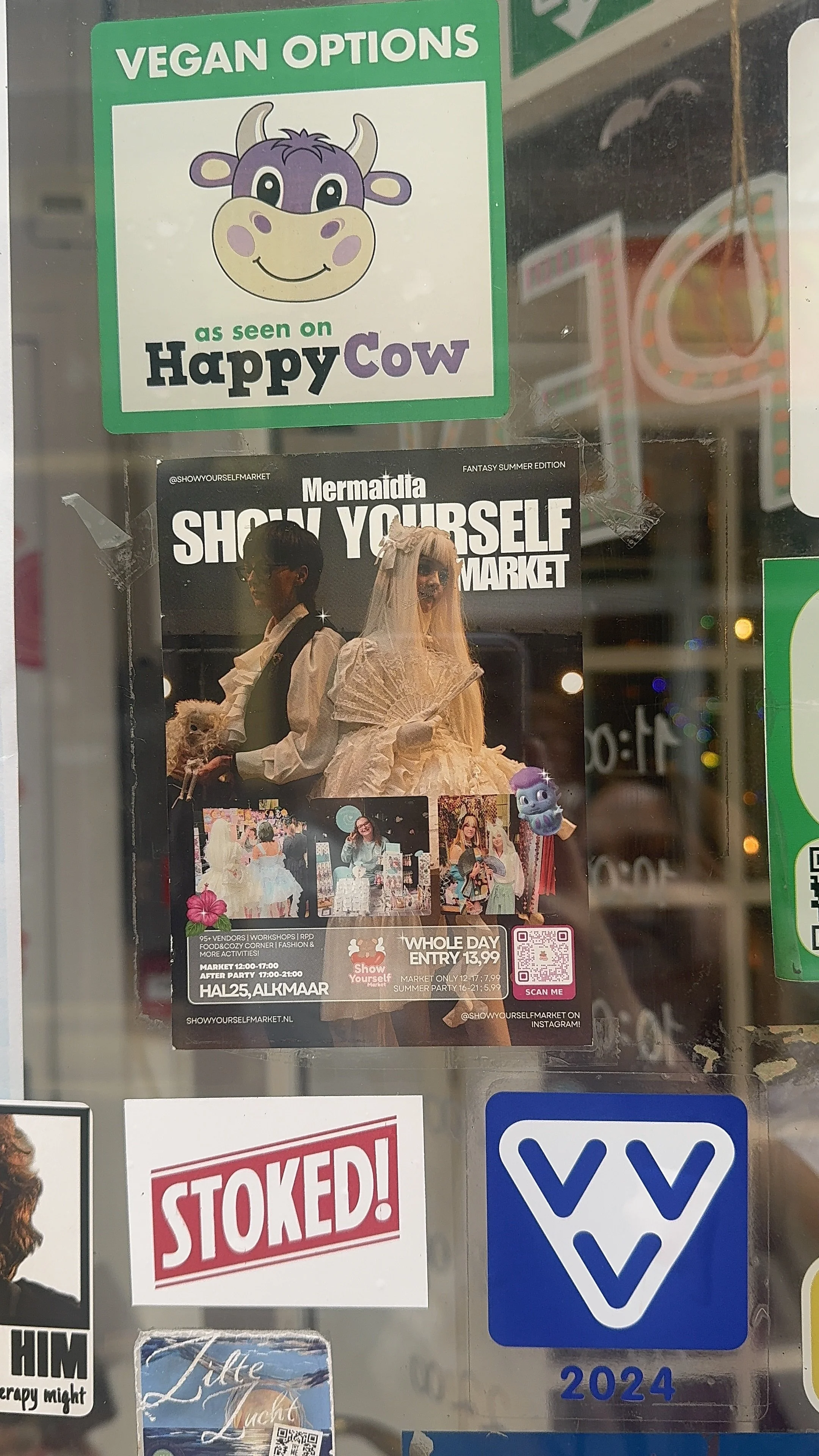 Een poster voor een markt met een afbeelding van mensen in kostuums en een cartoonachtig wezen. Bovenaan een groene sticker met de tekst 'Vegan Options' en een vrolijk koe-illustratie met de tekst 'as seen on Happy Cow'.