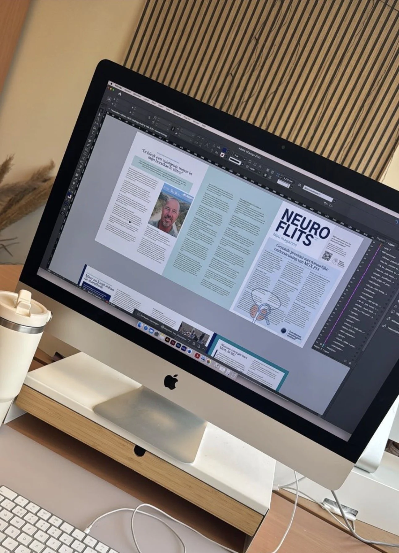 Een iMac-computer met een geopende grafische ontwerpsoftware die een gedrukte nieuwsbrief of magazine bewerkt, geplaatst op een wit bureau met een witte toetsenbord en een koffiebeker, op de achtergrond een houten of lamellen wand.