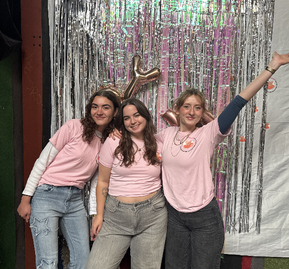 Drie jonge vrouwen glimlachen en poseren voor een feestje, met metallic slierten en ballonfiguren op de achtergrond.