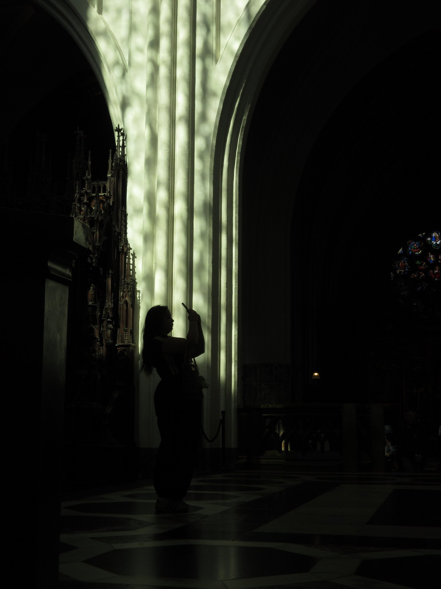 Een silhouet van een persoon die staat en een smartphone vasthoudt binnen in een kathedraal of kerk, met een groot raam en gekleurde raamstijlen op de achtergrond.