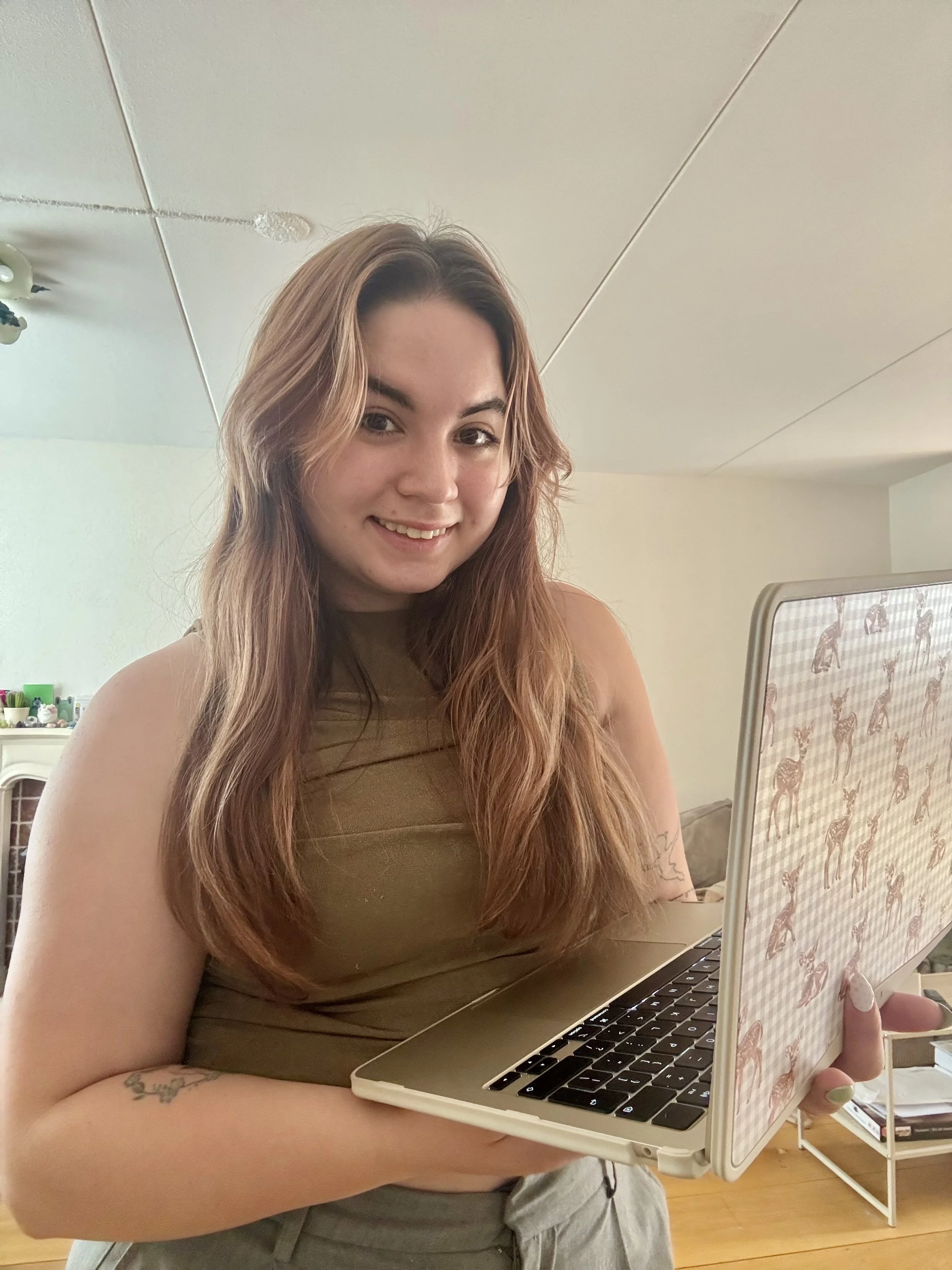 Een vrouw met lang, lichtbruin haar en een tatoeage op haar arm houdt een laptop met een hoesje met een giraffenpatroon. Ze zit in een kamer met een houten vloer en witte muren. In de achtergrond is een witte boekenplank met boeken en decoraties.