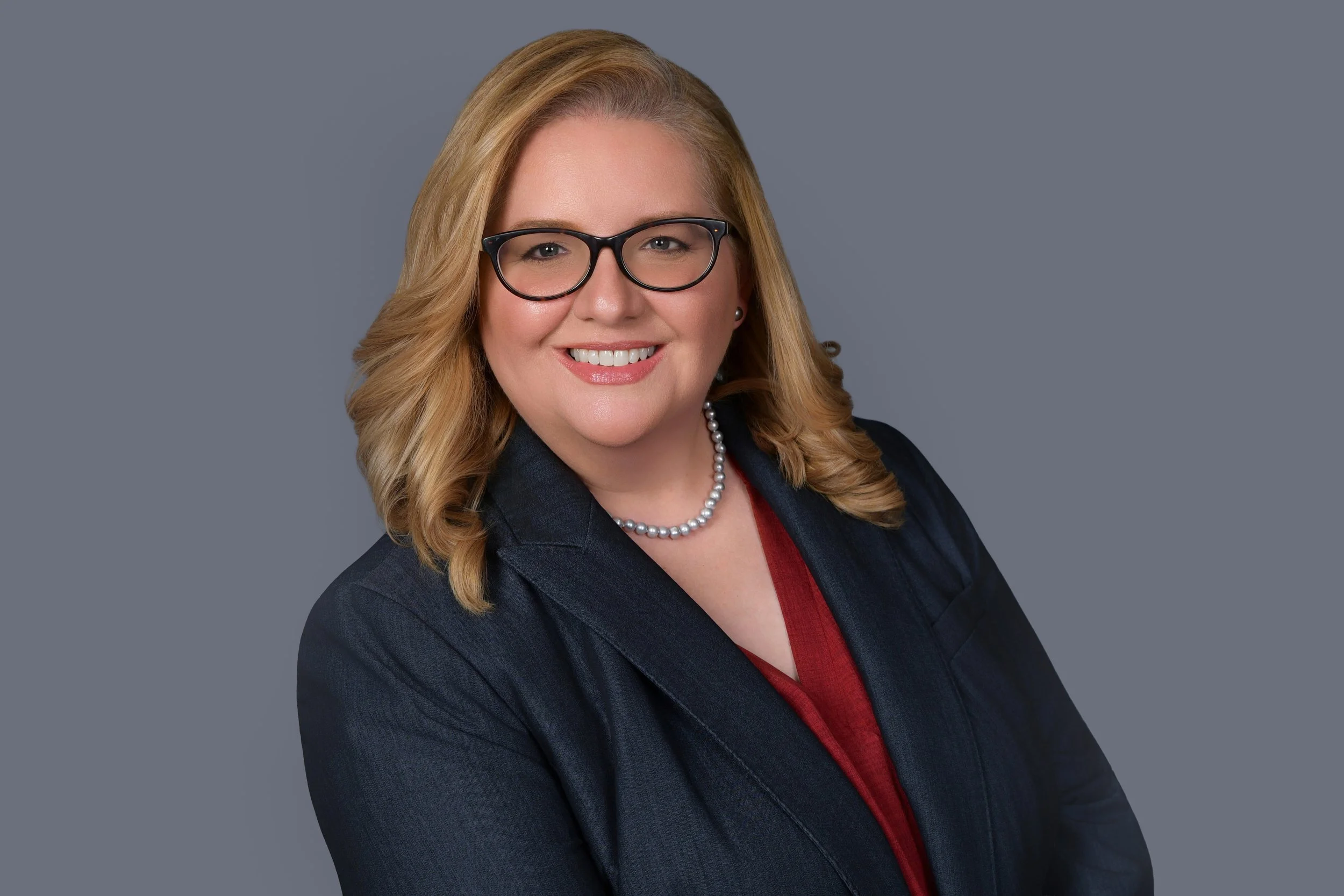 A headshot of a woman with blonde hair, wearing glasses, a dark blazer, a red blouse, and a pearl necklace, against a gray background.