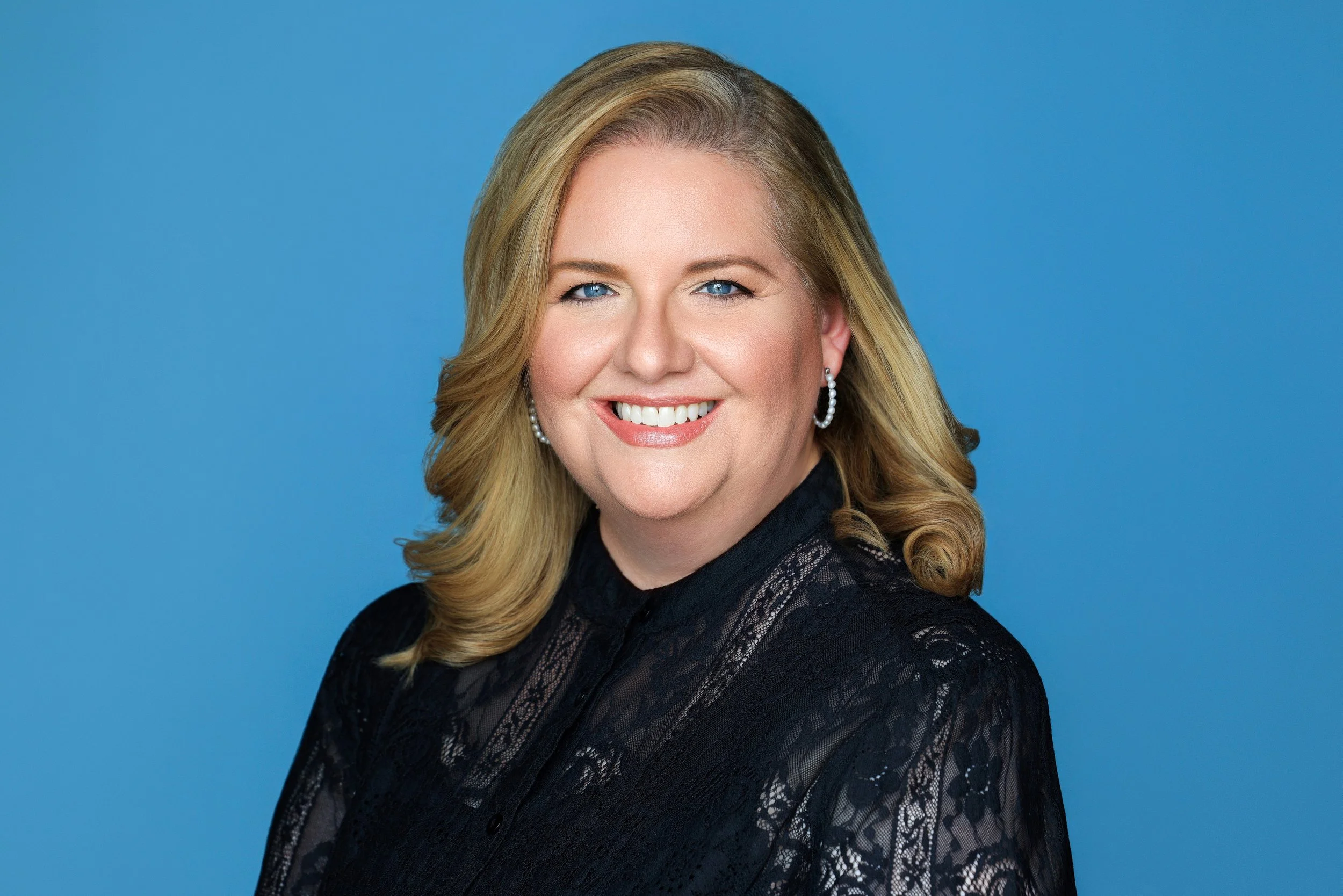 Portrait of a smiling blonde woman with blue eyes, wearing pearl earrings and a black lace top, against a blue background.