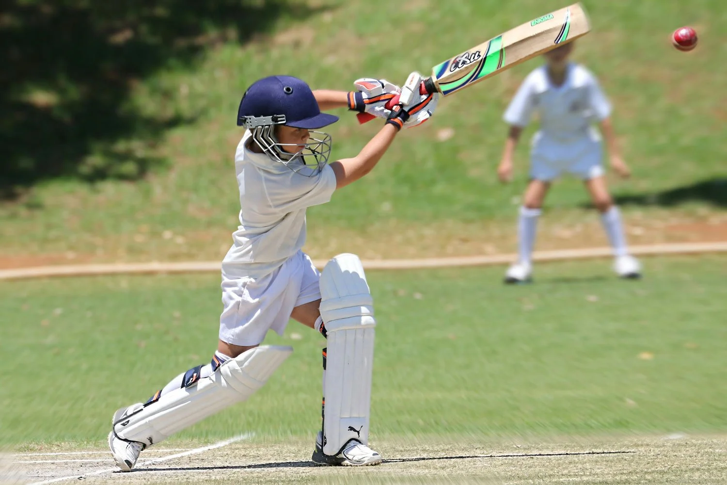 A young cricket player in white uniform, helmet, and pads, is hitting a cricket ball with a bat during a game.