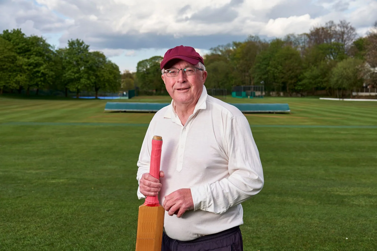 An elderly man wearing glasses, a white long-sleeve shirt, and a maroon cap, holding a wooden cricket bat wrapped in red tape on a grassy cricket field with trees and a cloudy sky in the background.