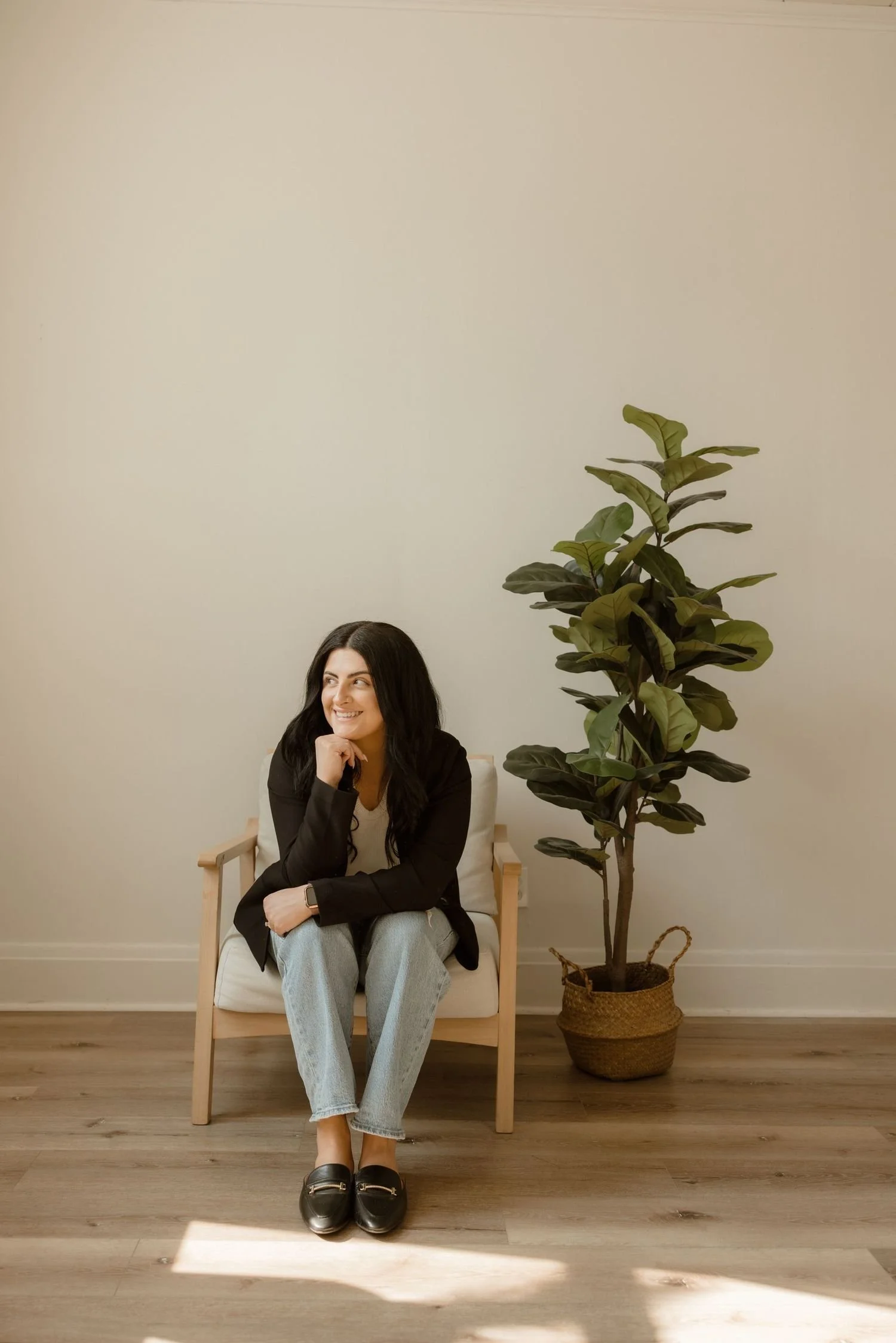 A woman with long black hair sitting on a white armchair with wooden armrests, smiling and resting her chin on her hand. She is wearing a black jacket, light blue jeans, and black loafers. There is a tall potted plant with dark green leaves next to her, and the background is a beige wall with wooden flooring.