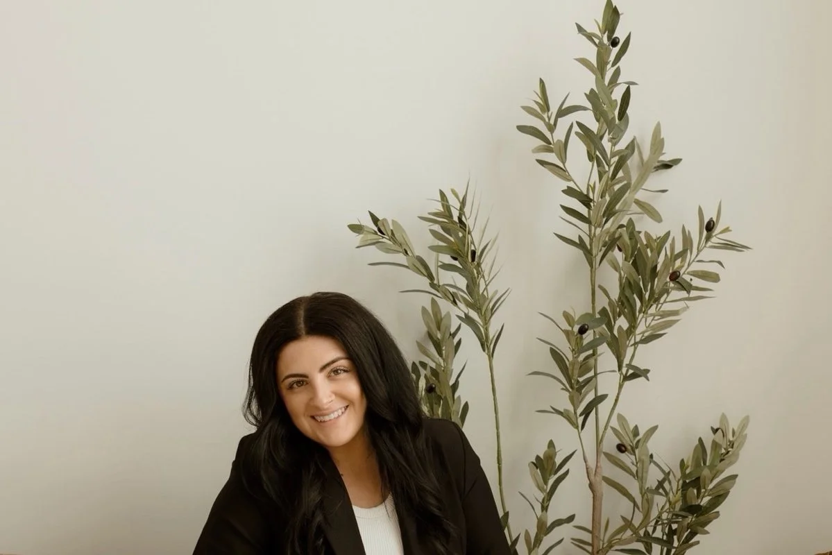 A woman with long dark hair smiling while sitting in front of a light-colored wall