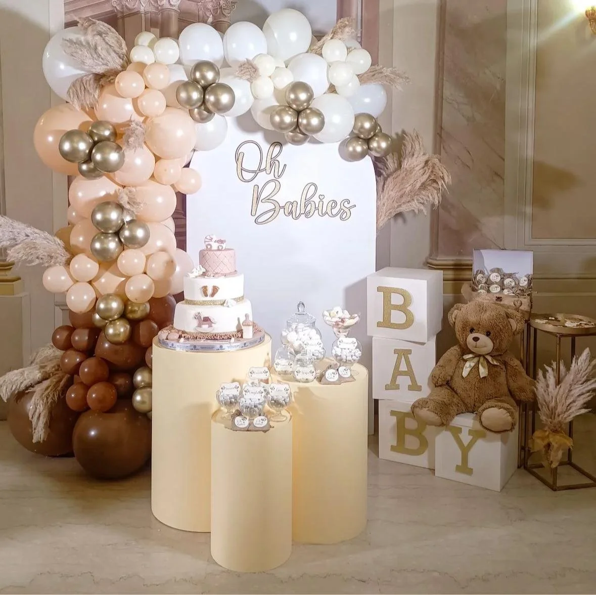 Decorazione per festa di nascita con palloncini bianchi, oro e marroni, torta, teddy bear e blocchi con scritte BA BY, sotto uno sfondo con scritta 'Oh Babies'.
