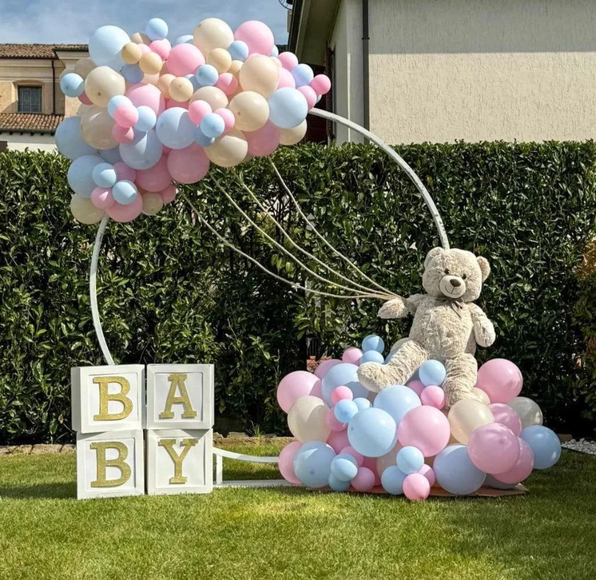 Decorazione per nascita con un orsetto di peluche seduto tra palloncini rosa, azzurri e crema, e lettere che formano la parola 'BABY' in blocchi bianchi