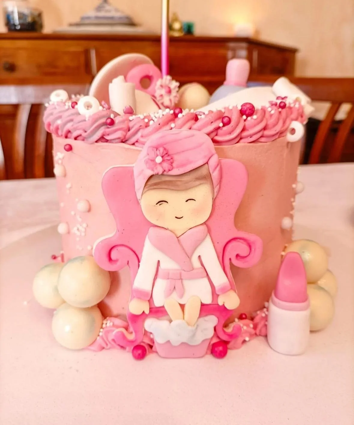 Torta decorata a tema bambina con figura di una ragazza in vestito rosa, ciocca di capelli, e accanto rossetto di cibo, con decorazioni di panna, palline di zucchero e confetti rosa e bianchi.