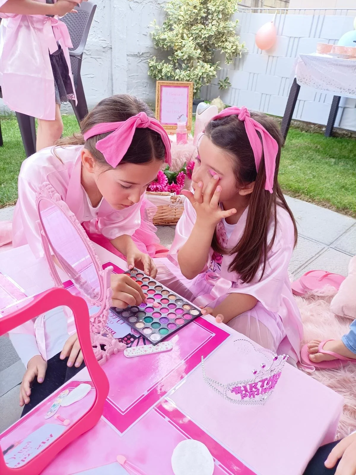 Due bambine celebrano un compleanno facendo trucchi sul viso, con decorazioni rosa e una corona di fronte a loro.