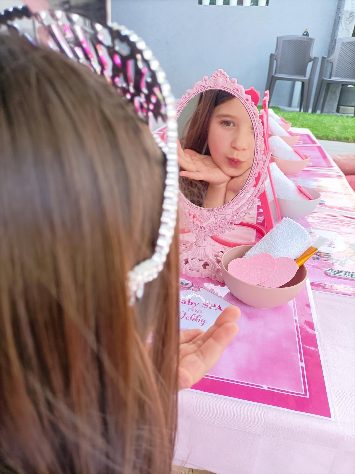 Una bambina con una fascia decorativa si guarda allo specchio; la scena è decorata con rosa e contiene accessori per il taglio di capelli o il trucco, tra cui un tovagliolo e una spatola.