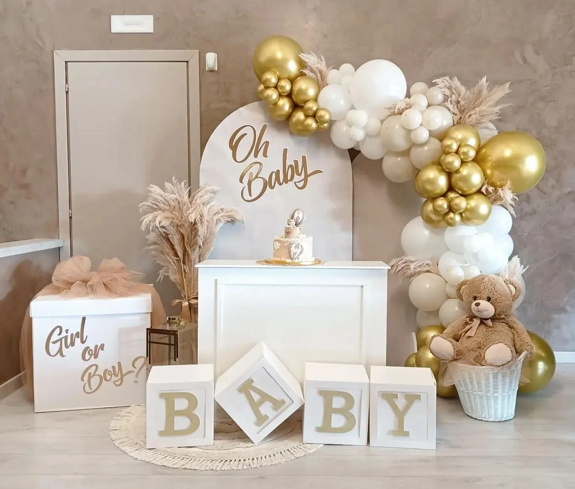 Decorazioni per una festa di nascita con palloncini bianchi e dorati, un grande cartellone con scritto Oh Baby, scatole con lettere B A B Y, teddy bear seduto in una cesta e una torta con decorazioni rosa.