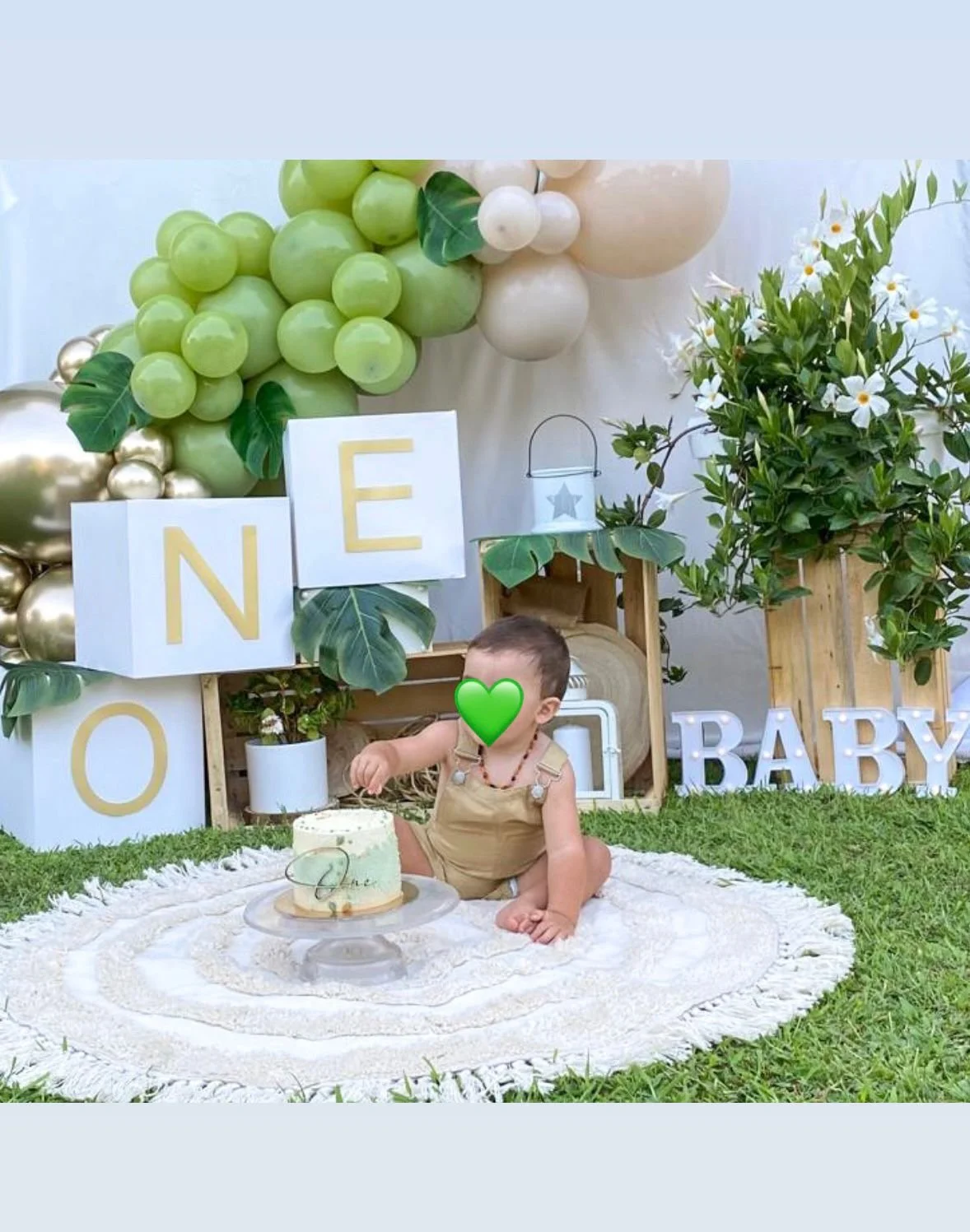 Bambino che si siede su un tappeto bianco con una torta, in uno sfondo decorato con palloncini e lettere che formano 'NENO BABY'.