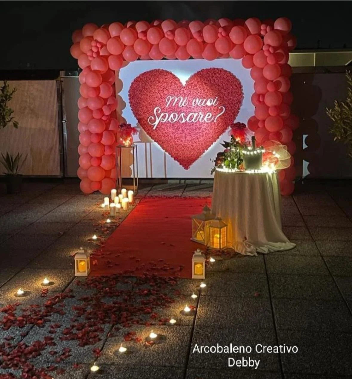 Decorazione romantica con cuore di fiori e palloncini rosa, con la scritta 'Mi vuoi sposare?' in primo piano, camino di candele e un tavolo con fiori e luci, in un'ambientazione serale.