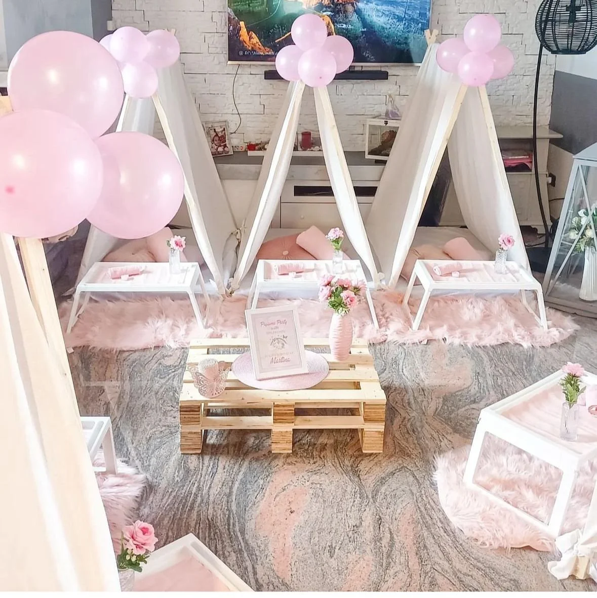 Decorazione per festa con tende bianche, palloncini rosa, tavolini bianchi con fiori, cuscini rosa, tappeti rosa e vassoi con dettagli rosa.
