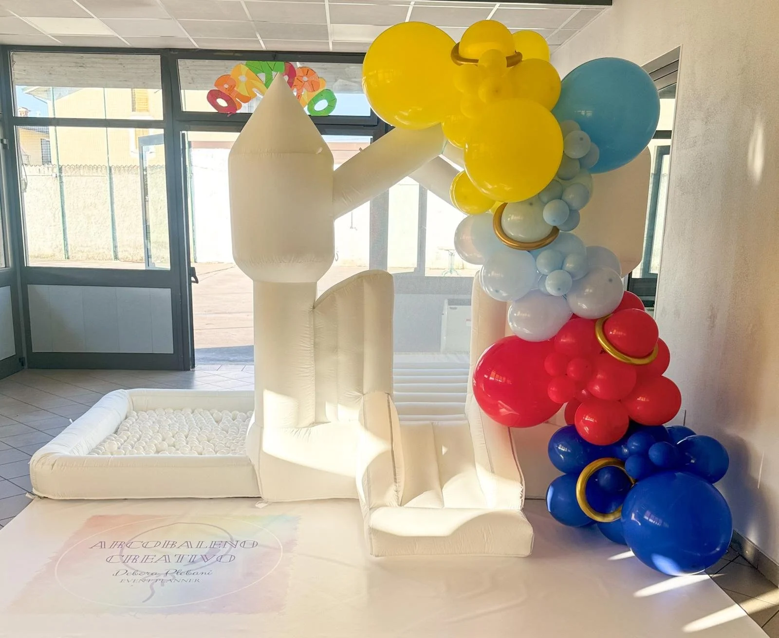 Decorazione con palloncini colorati e un grande lettera illuminata in forma di L, situata in un locale con ingresso e pareti di vetro, probabilmente per un evento o festa.