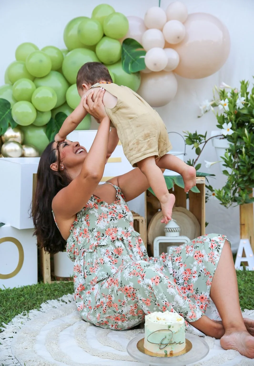 Una donna seduta sul tappeto sorride mentre solleva un bambino in aria in un giardino decorato con decorazioni di frutta artistica, tra cui uva e meloni, e un dolce sulla tazza.