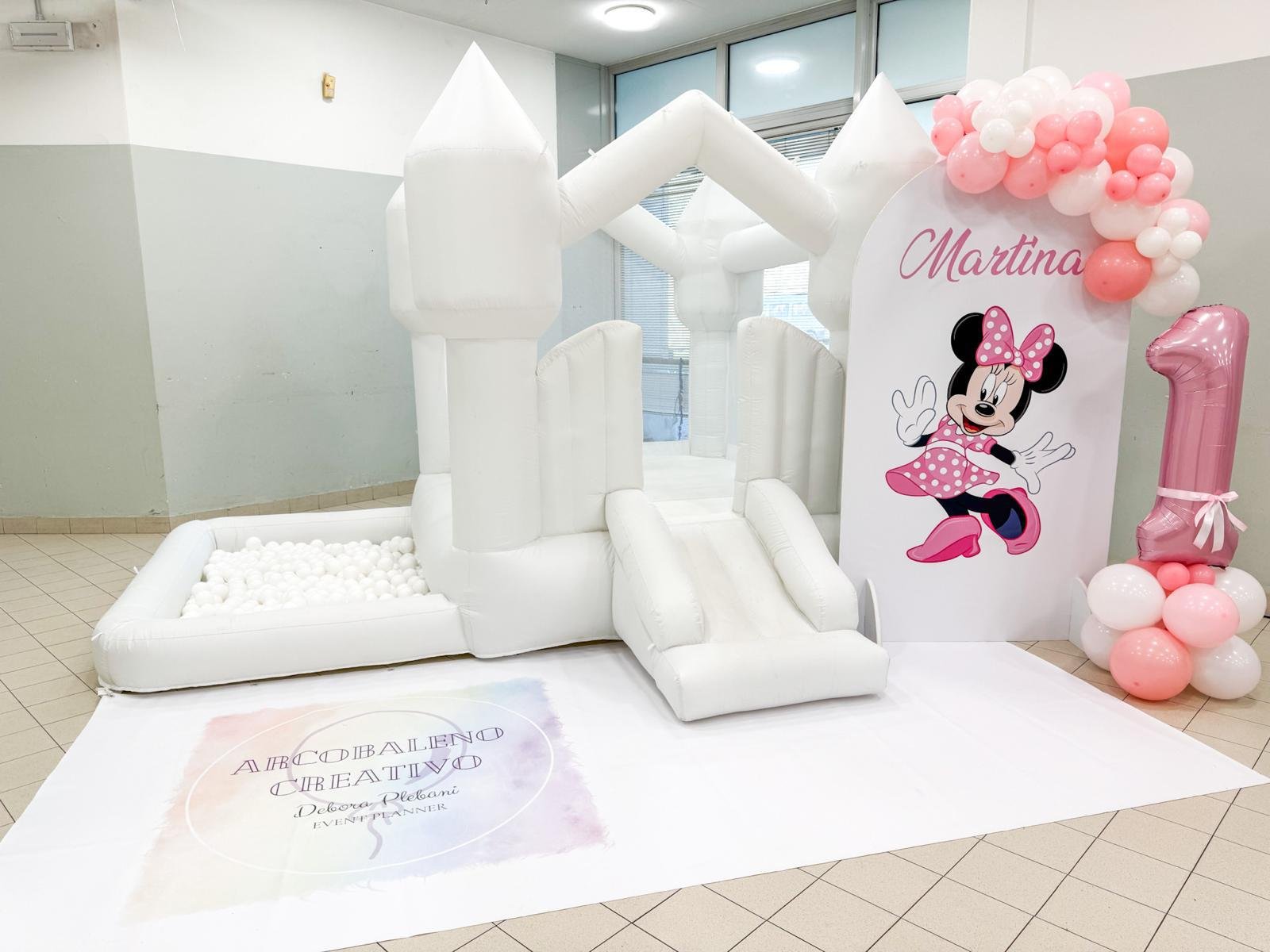 Decorazione di compleanno con castello gonfiabile bianco, un grande cartellone con Minnie Mouse, palloncini rosa e bianchi con il numero uno, e un tappeto con scritte. Il cartellone dice 'Martina' e mostra Minnie Mouse in posa.