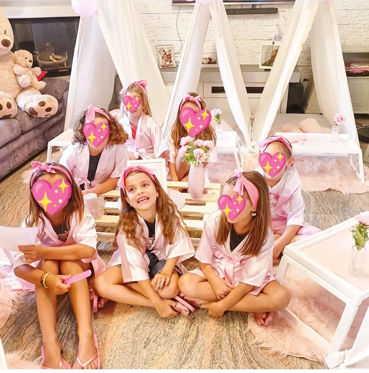 Gruppo di bambine sedute in una stanza decorata con tende bianche, fiori e mobili rosa, tutte indossano pigiami rosa e copricapi rosa. Alcune sono sedute a terra, altre su sedie o su un banco.