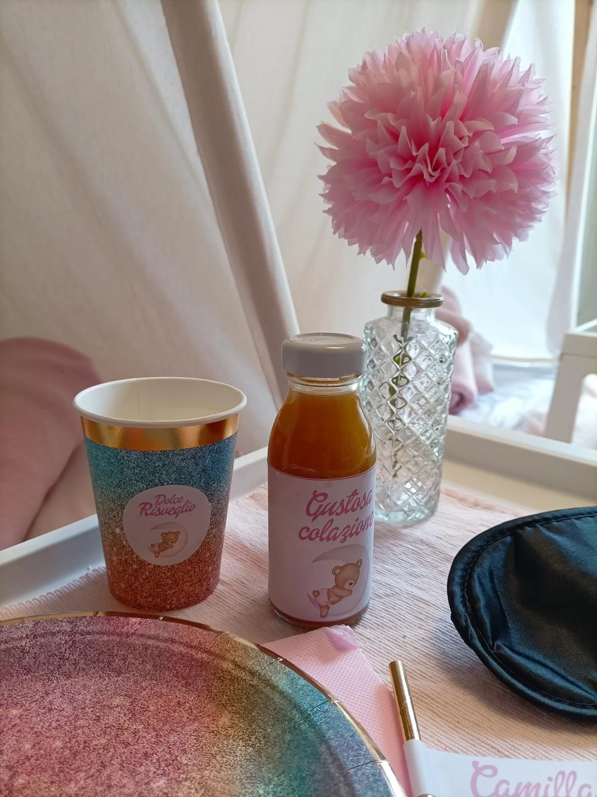 Bottiglia di succo con etichetta rosa e disegno di un orsetto, bicchiere con decorazioni colorate, posate, piatto, e fiore rosa in un vaso di vetro cristallino su una tovaglia rosa