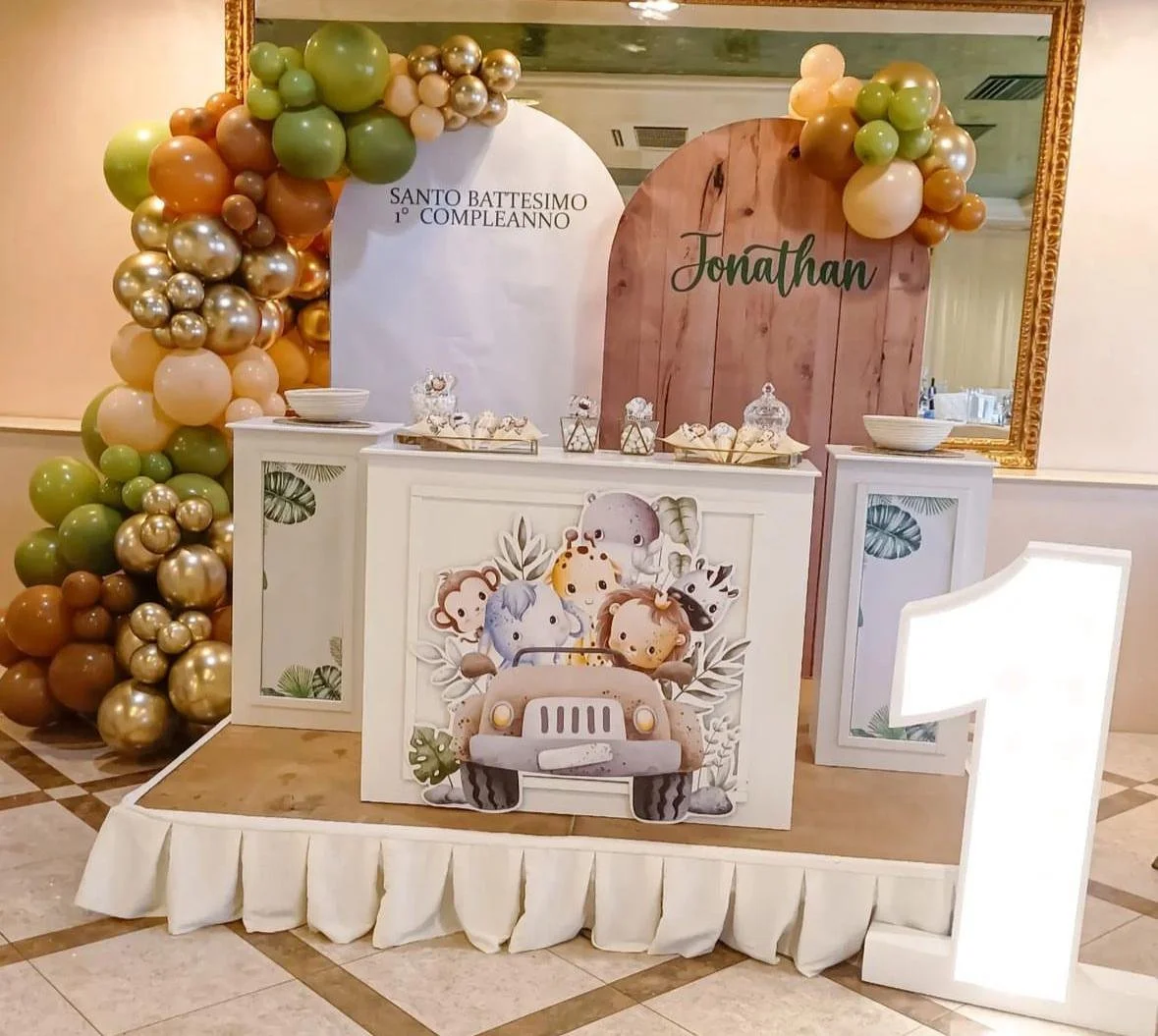 Decorazione per battesimo e primo compleanno con palloncini dorati, verdi e bianchi, e un pannello con il nome Jonathan, con un'illustrazione di animali in un'auto. La scena include tavoli con dolci e un grande numero 1 illuminato.