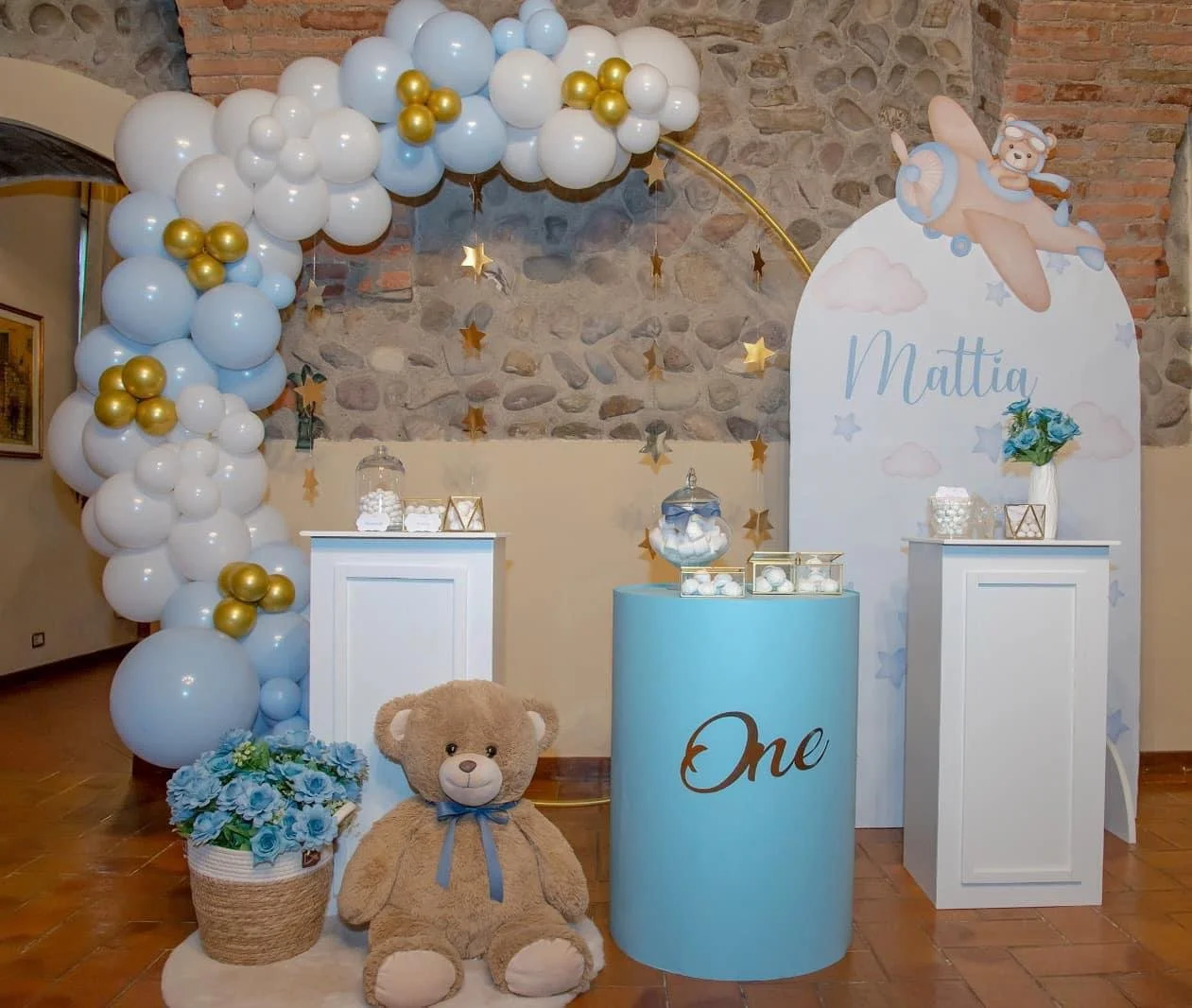 Decorazioni di festa per bambino con palloncini bianco e oro, un grande cartellone con il nome Mattia, un orsetto di pezza, una scatola con confetti, fiori blu e dolci. Atmosfera festiva e dolce.
