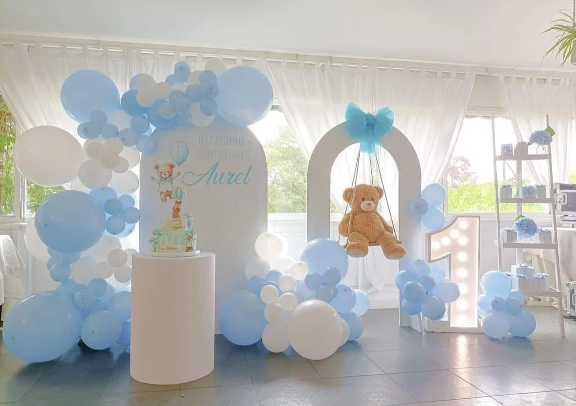 Decorazioni per festa di compleanno di Aurel, con palloncini azzurri e bianchi, un grande numero 1 illuminato, un orsetto di peluche e un pannello con torta e slogan in italiano.