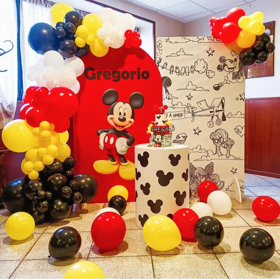 Decorazioni di compleanno a tema Mickey Mouse con palloncini rossi, gialli, neri e bianco, un grande cartellone con immagine di Mickey Mouse e il nome Gregorio, e una torta decorata con Mickey Mouse e palloncini.