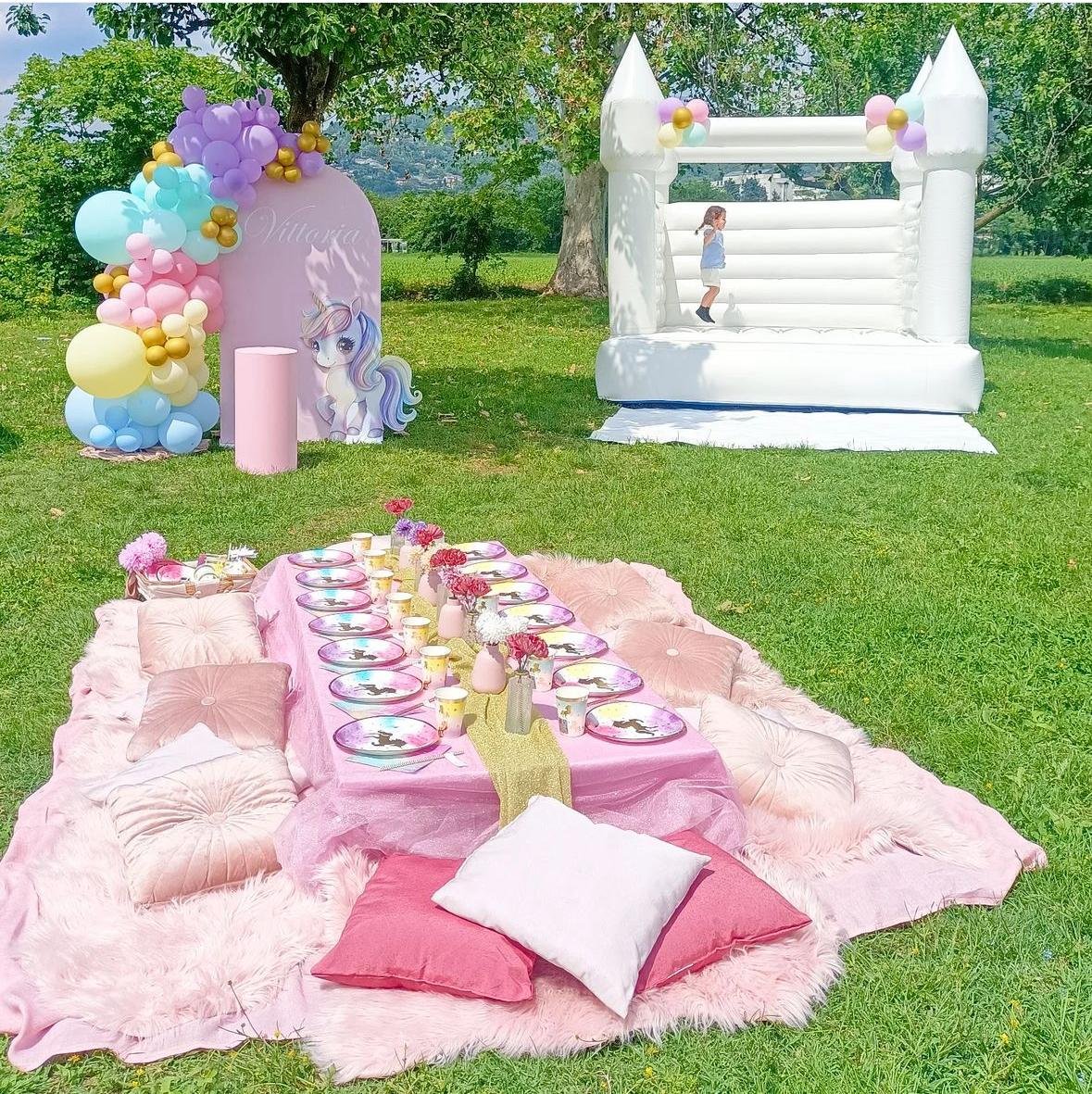 Decorazione per festa di compleanno a tema unicorni, con un castello gonfiabile bianco, un tavolo con piatti e bicchieri decorati, e un'area di seduta con cuscini e tappeti rosa.