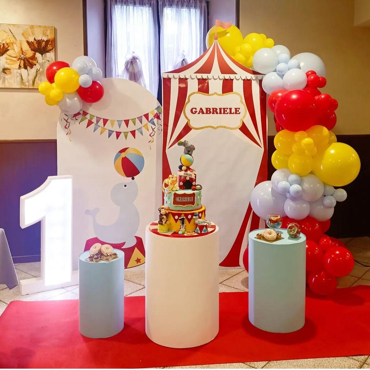 Decorazioni per festa di compleanno con tema circus, including palloncini rossi, gialli e bianchi, un grande numero 1, una torta decorata e uno sfondo a strisce rosse e bianche con il nome Gabriele.