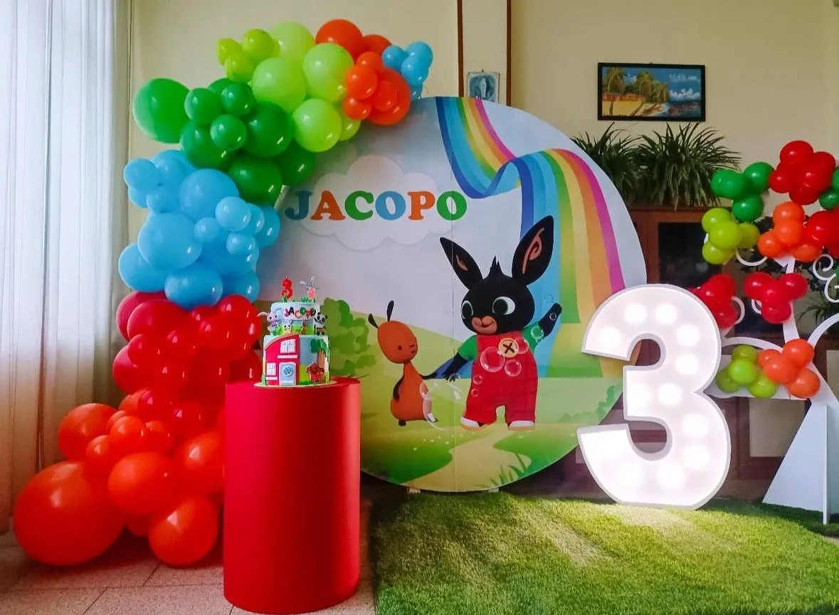Decorazione di compleanno per un bambino con palloncini colorati, una torta in stile cartone animato e una grande lettera '3' luminosa.