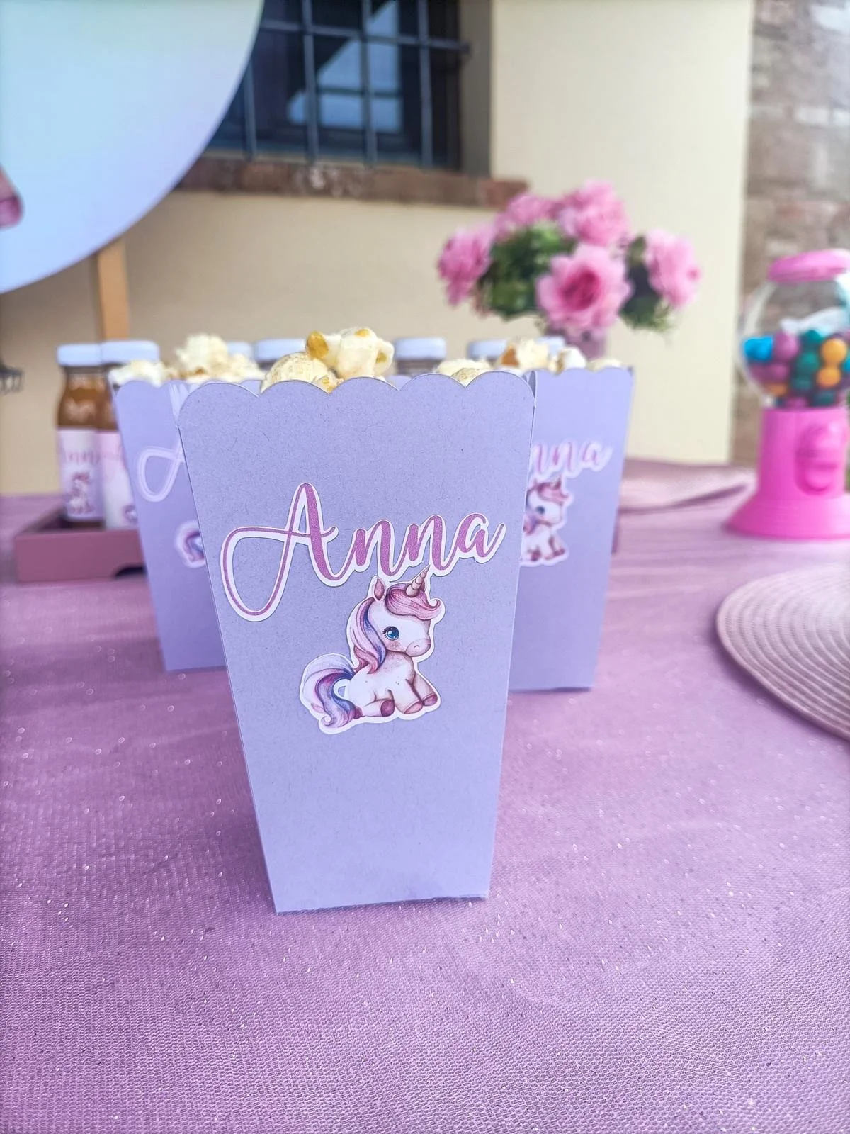 Decorazioni per una festa di compleanno con etichette a forma di barattoli decorati con un unicorno e il nome Anna, fiori rosa sullo sfondo e una macchina dei gumbi.