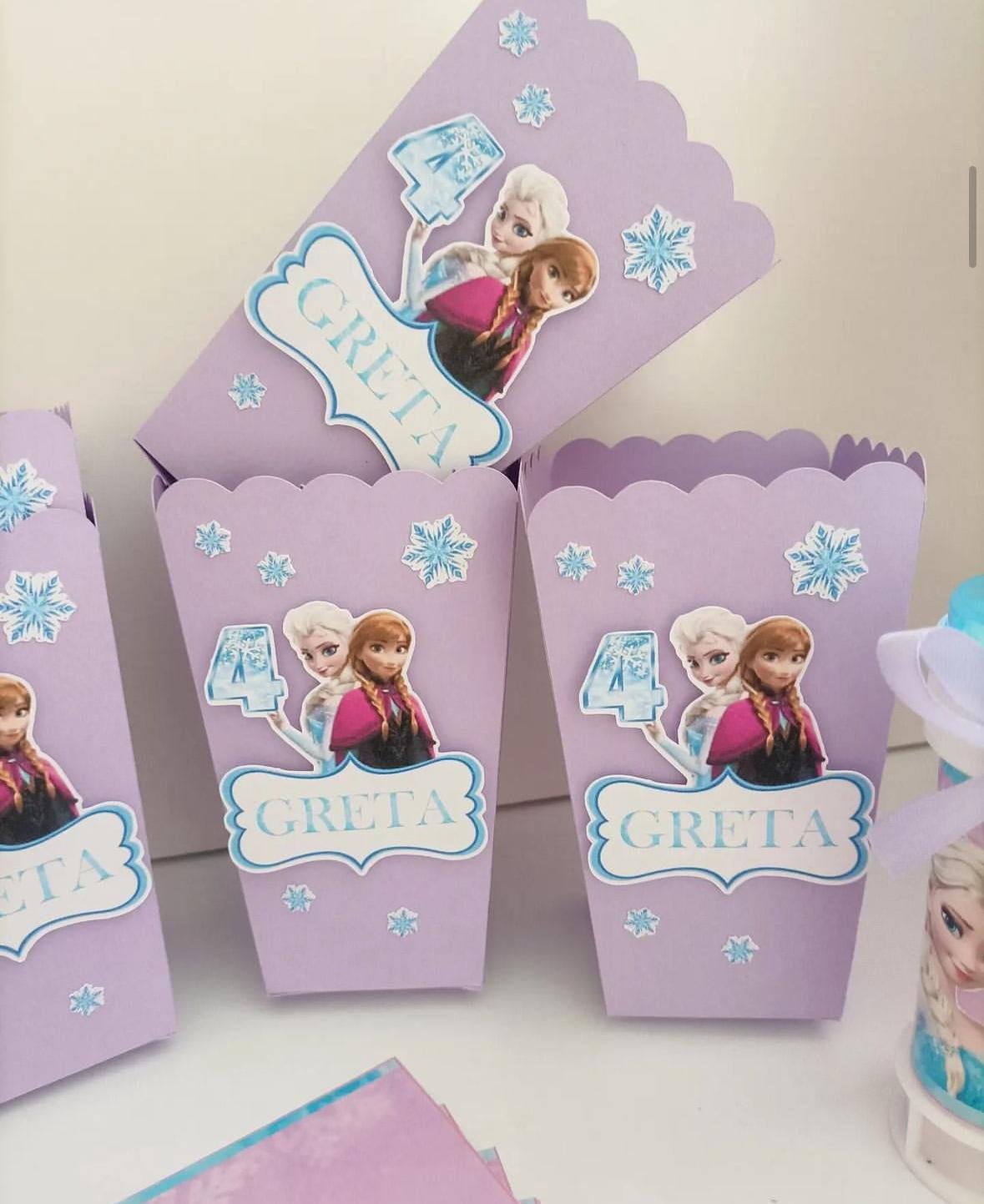 Confezioni di cartone color lilla con decorazioni di fiocchi di neve e adesivi di Elsa e Anna di Frozen. Ogni confezione ha il numero 4 e il nome Greta.