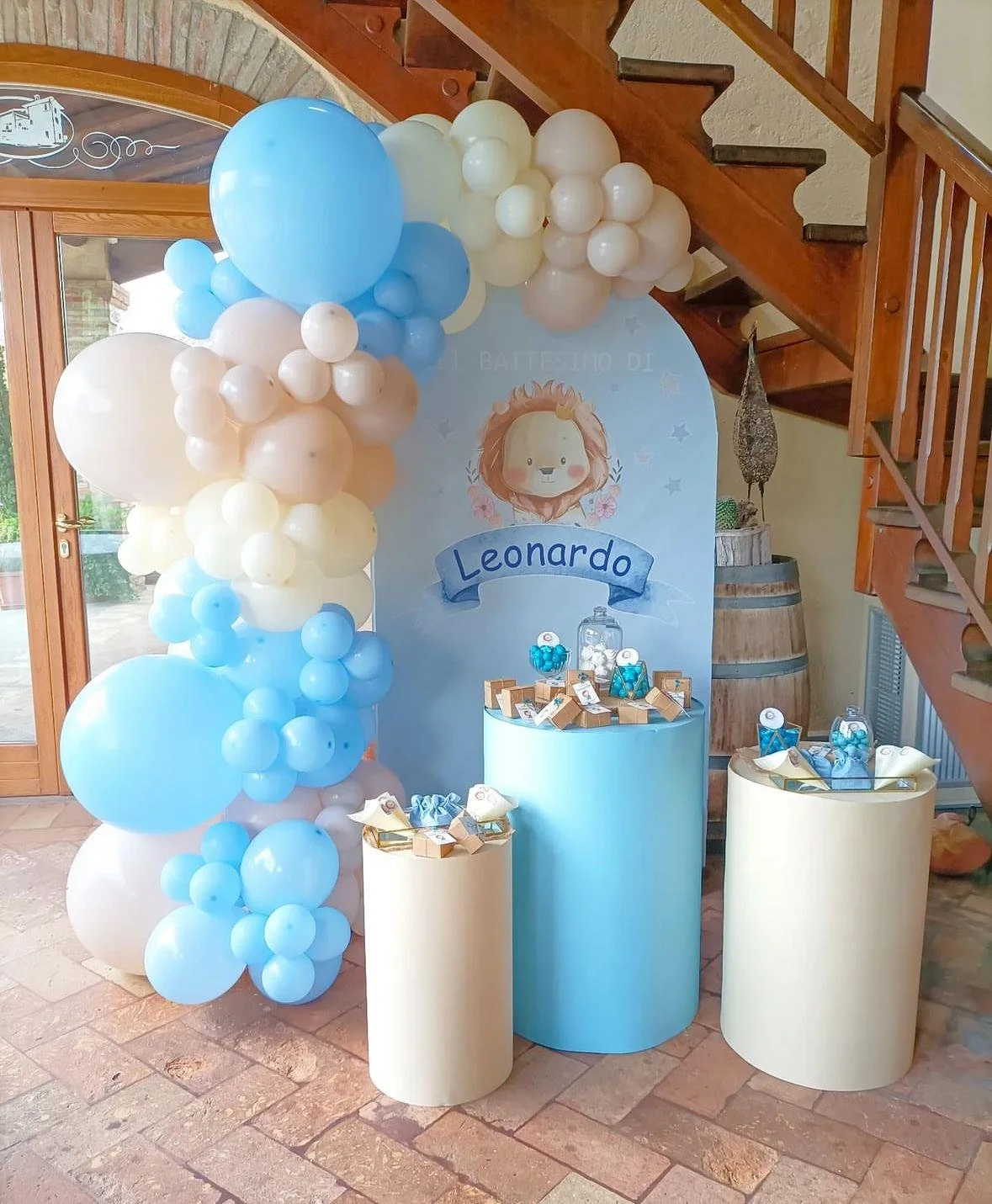 Decorazioni per battesimo di Leonardo con palloncini azzurri, bianchi e beige, cornice con disegno di leone e nome Leonardo, tavoli con dolci e regali.