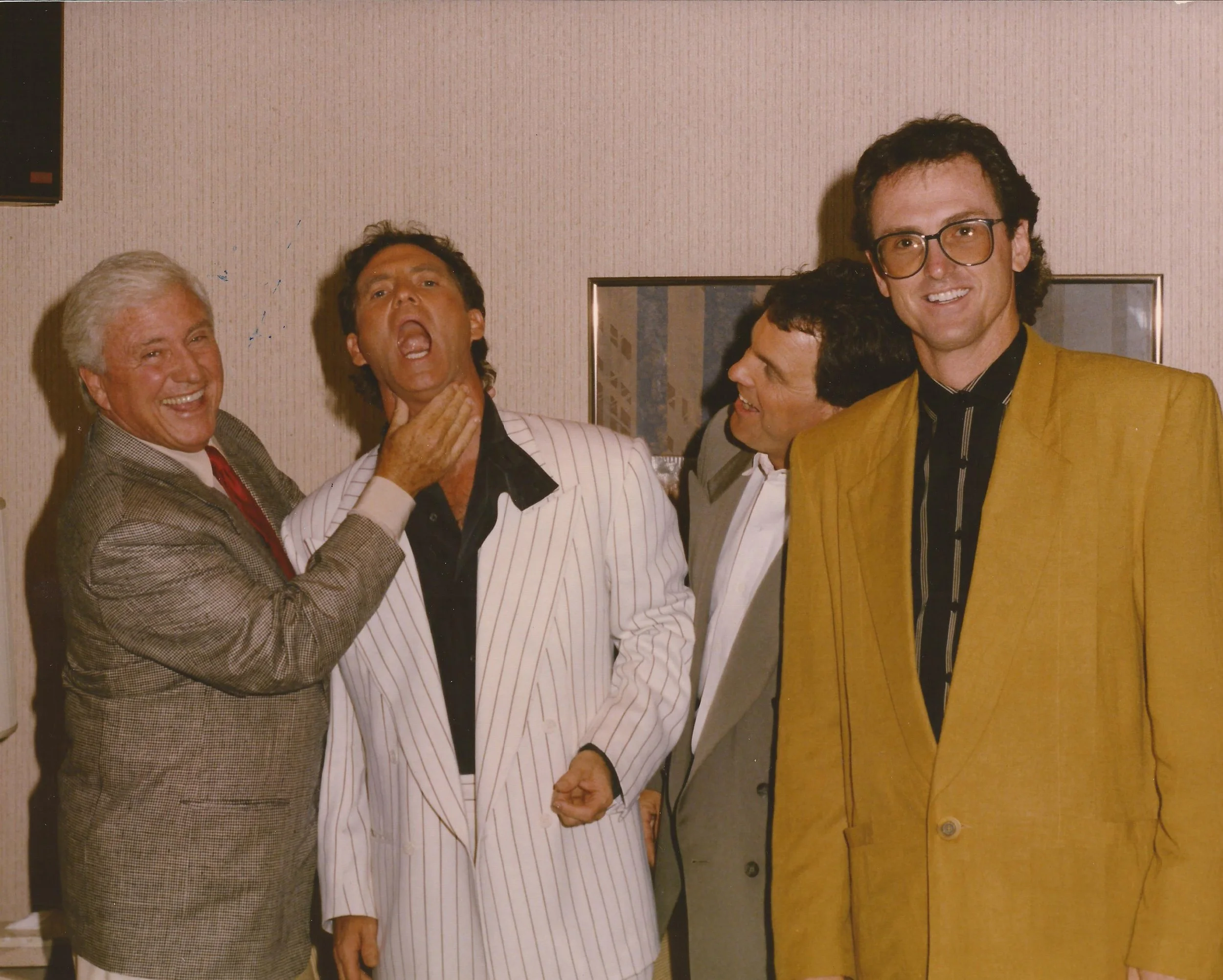 Four men are standing together in a room, smiling and posing for the photo. The man on the far left has gray hair and is wearing a gray patterned suit with a white shirt and red tie. The second man from the left has dark hair and is wearing a white pinstripe suit with a black shirt. The third man, partly obscured, has dark hair and is wearing glasses and a white shirt. The man on the far right has dark, curly hair, glasses, and is wearing a yellow blazer with a black shirt that has gold and white stripes.