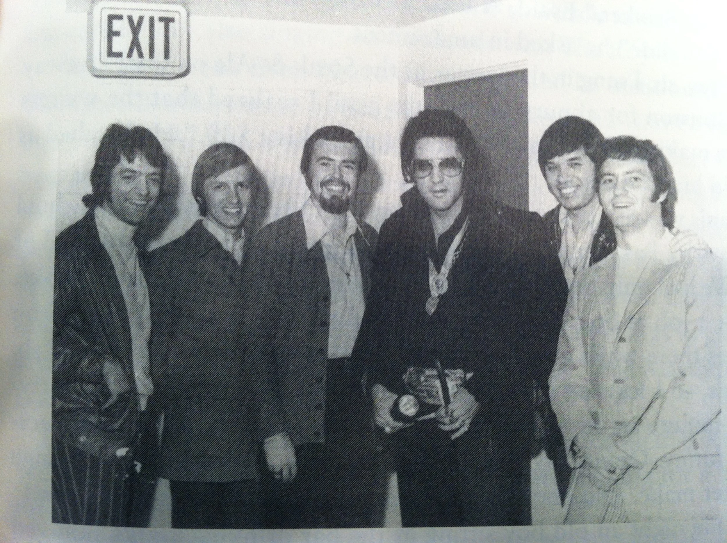 Black and white photo of six young men standing close together, smiling, inside a building near an exit sign.