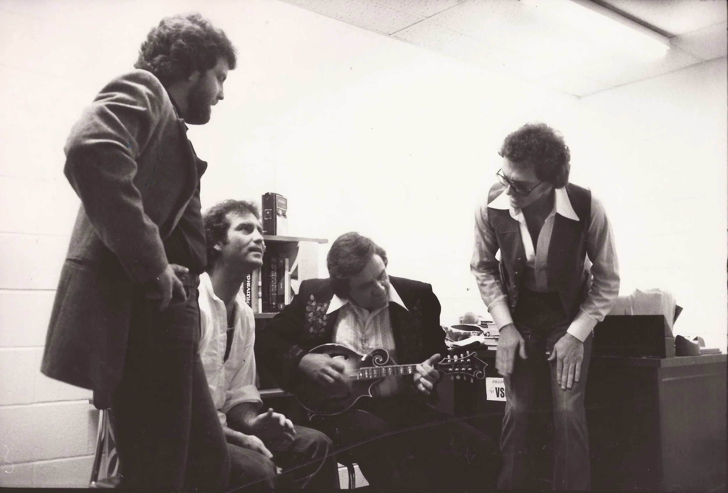 Four men in a room, two of them are sitting and two are standing, with one playing guitar and others listening.