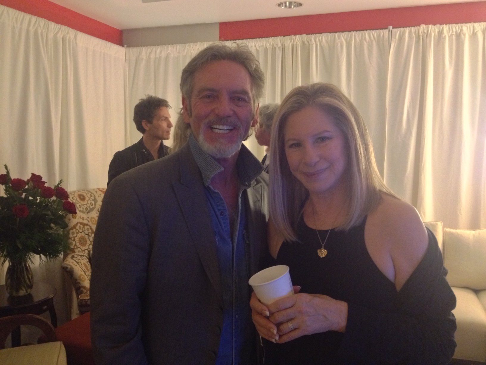 A man with gray hair and beard, dressed in a gray suit, smiling next to a woman with blonde hair, wearing a black dress and heart-shaped necklace, holding a paper cup, at a social gathering with cream curtains and floral wallpaper.