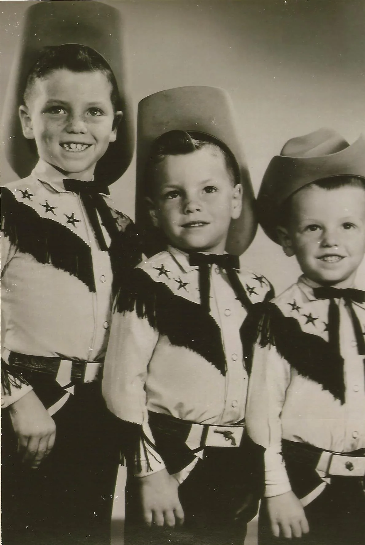 Three young boys dressed in cowboy costumes standing in a line. They are wearing cowboy hats, shirts with star decorations, and fringed vests.