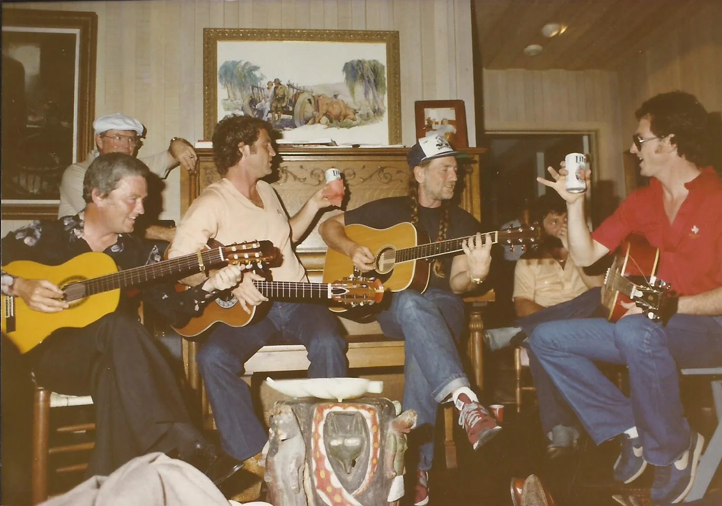 A group of men sitting and playing guitars, with some holding beer cans, in a cozy, decorated room with framed art on the walls.